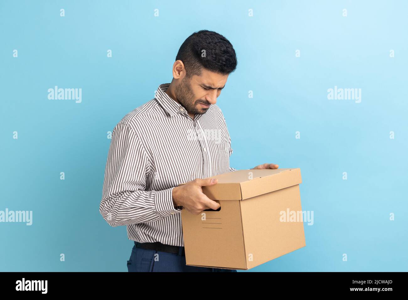 Portrait of sad worker man holding cardboard parcel, upset about being fired at work, crying, expressing sorrow, wearing striped shirt. Indoor studio shot isolated on blue background. Stock Photo