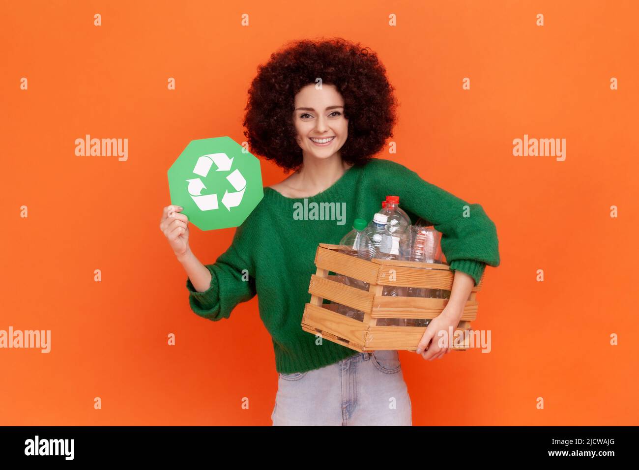 Smiling woman with Afro hairstyle wearing green casual style sweater holding box with plastic bottles and showing green recycling sign. Indoor studio shot isolated on orange background. Stock Photo