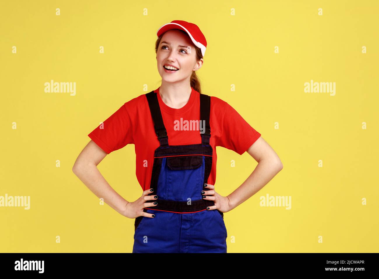 Portrait of positive dreamy worker woman standing with hands on hips, looking away, thinking about something pleasant, wearing overalls and red cap. Indoor studio shot isolated on yellow background. Stock Photo