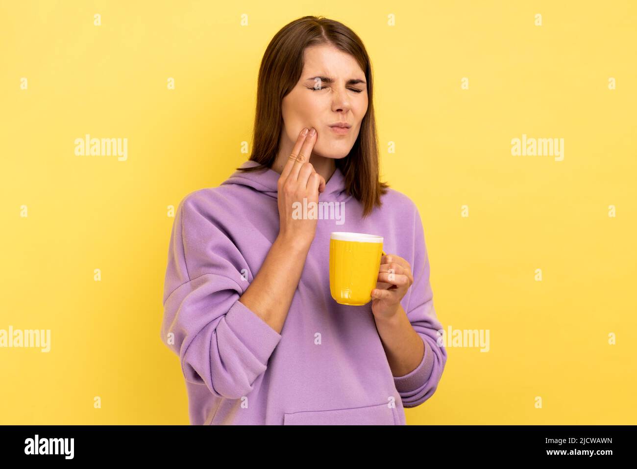 Portrait of sad sick unhealthy woman has teeth pain after drinking hot or cold beverage, dental injury, wearing purple hoodie. Indoor studio shot isolated on yellow background. Stock Photo