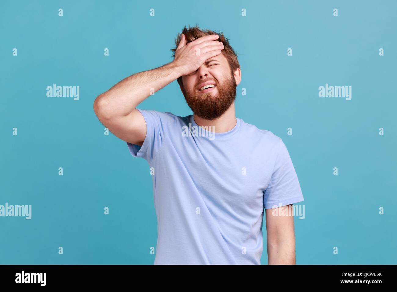 Portrait of upset sad bearded man standing with facepalm gesture, blaming himself, feeling sorrow regret because of bad memory. Indoor studio shot isolated on blue background. Stock Photo