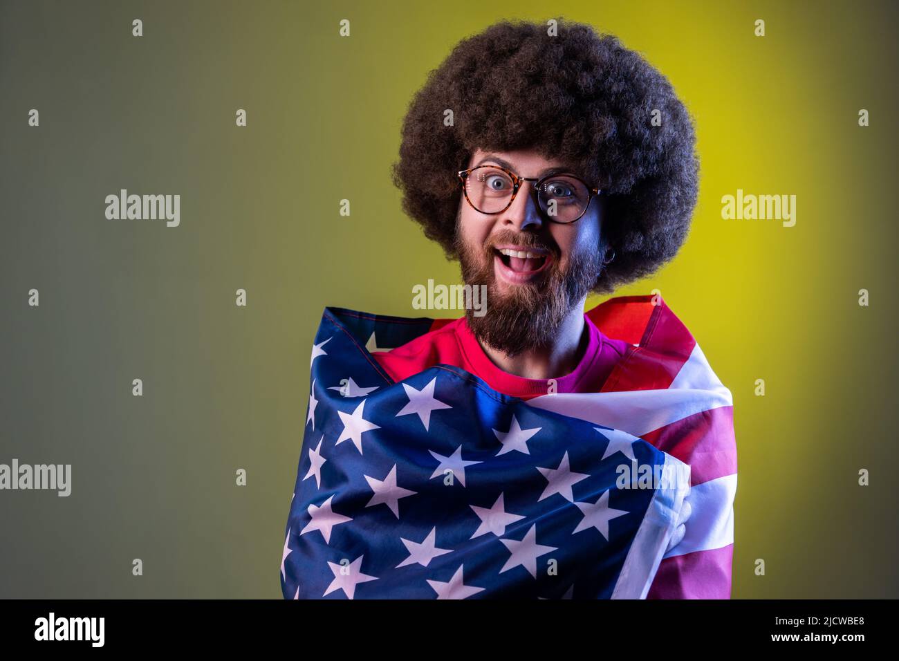 Portrait of hipster man standing wrapped in american flag, looking at camera with amazed excited facial expression. Indoor studio shot isolated on colorful neon light background. Stock Photo