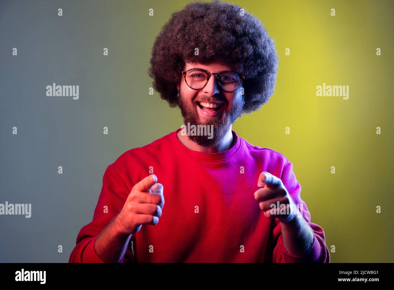 Portrait of hipster man with Afro hairstyle pointing to camera, smiling, making happy choice, we need you concept, wearing red sweatshirt. Indoor studio shot isolated on colorful neon light background Stock Photo