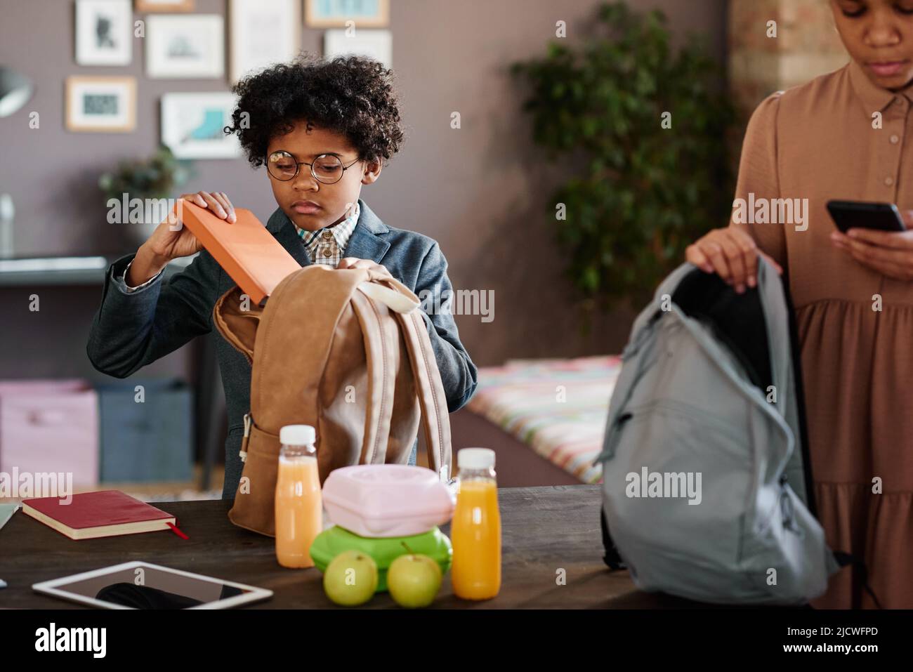 African little boy in eyeglasses packing books and lunch box in his school bag before going to school Stock Photo