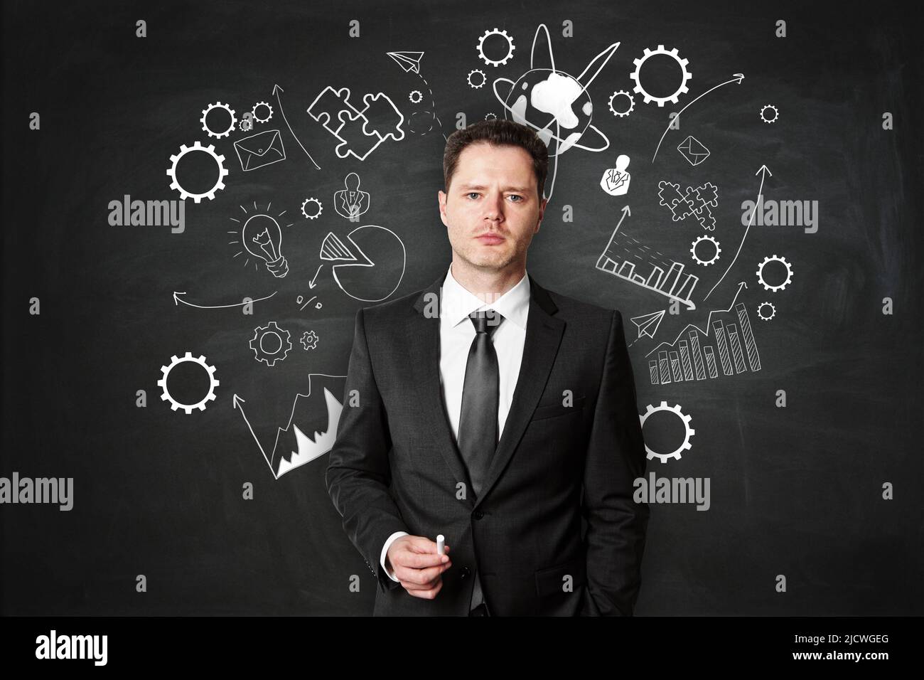 Business school education offline concept with teacher holding chalk in his hand on blackboard background with white handwritten puzzle, gears and arr Stock Photo