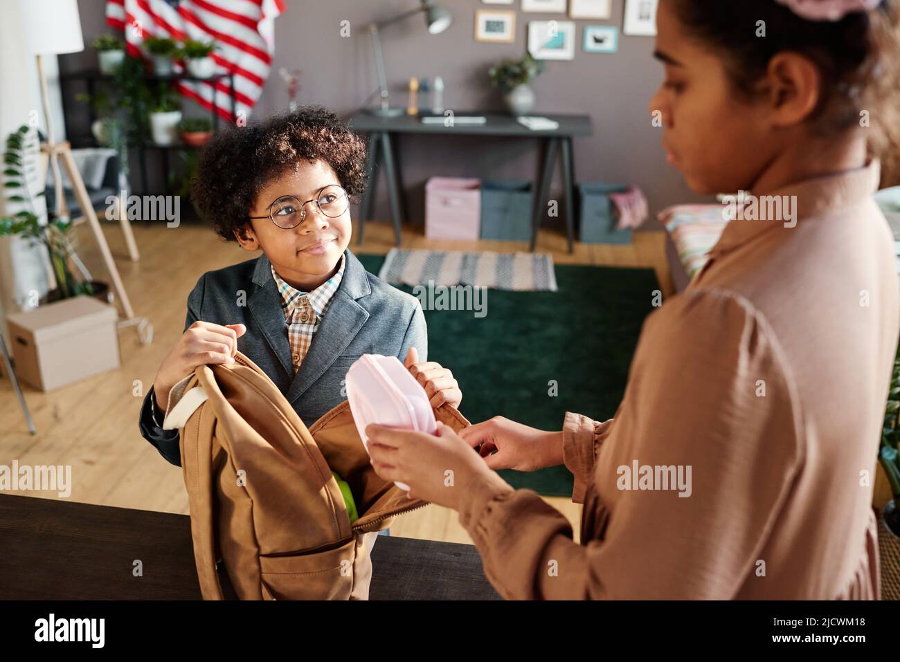 African little boy in eyeglasses opening his school bag while his elder sister packing lunch box in it and helping him Stock Photo