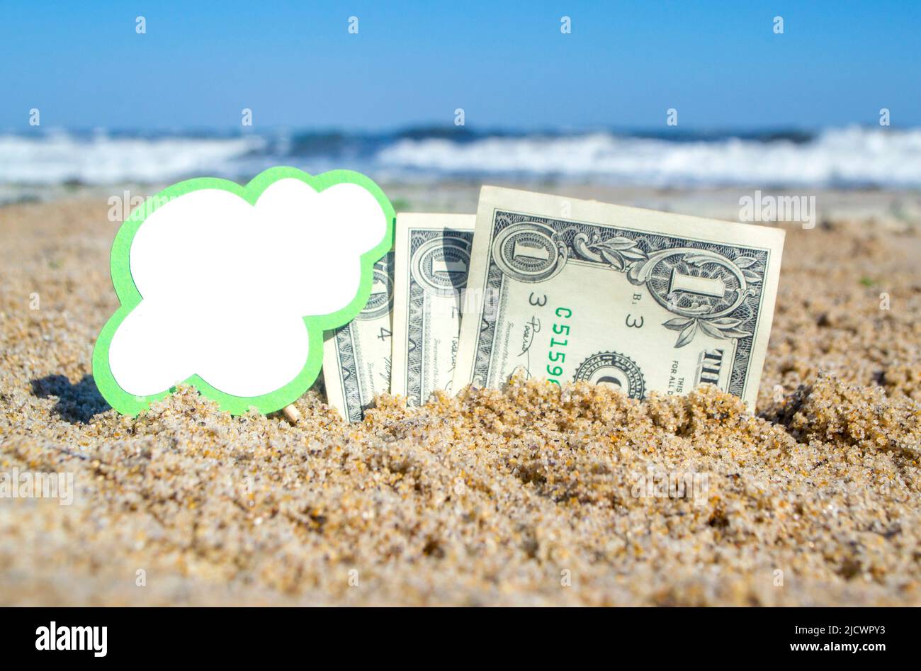 Paper bills one dollar and small green stick with clean empty paper speech bubble buried in sand beach background sea close-up in sunny summer day. Concept Money travel tourism vacation holiday relax Stock Photo