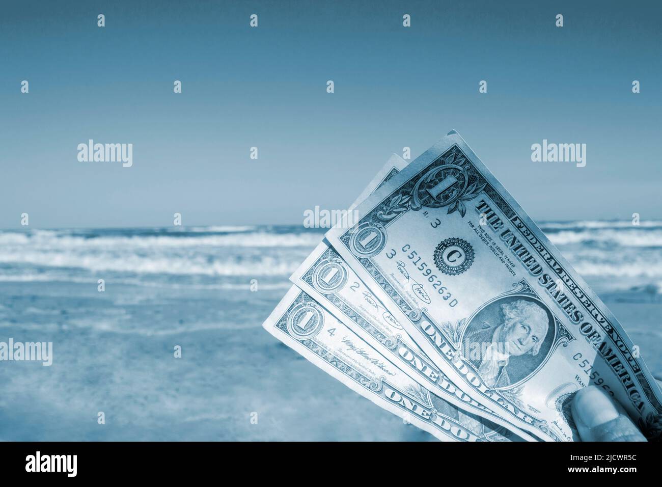 Person holding paper dollar bills on background of sea and sea waves, sandy beach and clear blue sky on sunny summer day. Concept of money, expenses on vacation, travel, tourism. Blue color Stock Photo