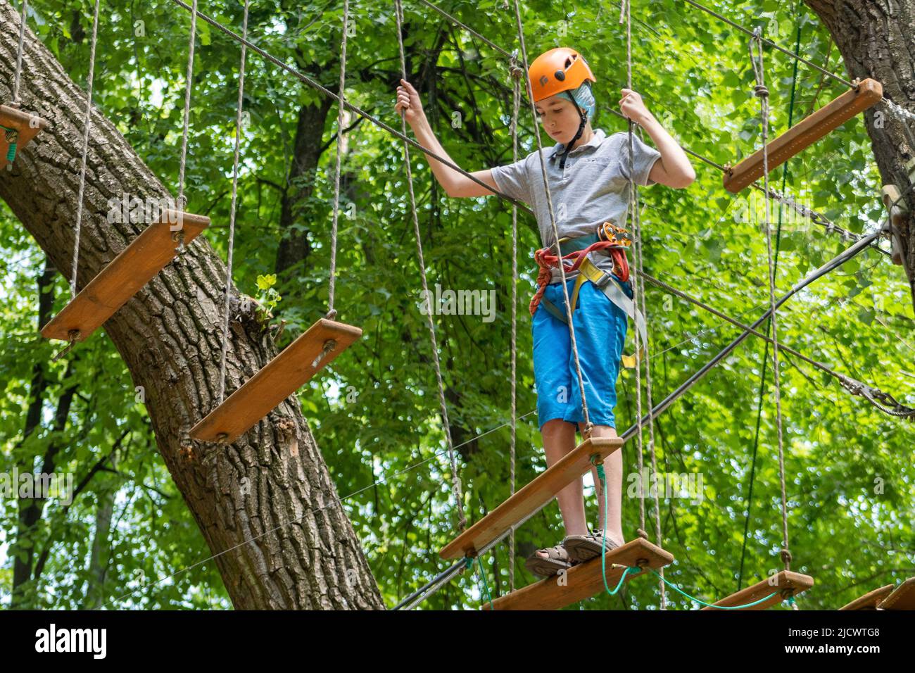 Rope park. A boy teenager in a helmet walks on suspended rope ladders. Carabiners and safety straps. Safety. Summer activity. Sport. Children's playground in nature in the forest. Stock Photo