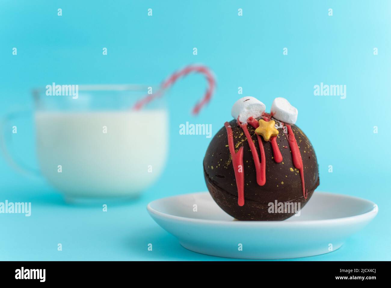 Chocolate bomb with marshmallows and Christmas decorations on a white plate against a blurred image of a glass of milk. blue background. High-quality photo Stock Photo