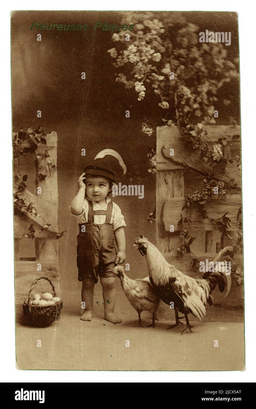Original kitsch Austrian published Easter greetings card postcard intended for the French market, depicting a cute little boy wearing traditional lederhosen (leather shorts) style shorts, hat with feather, hen & cockerel, basket of eggs, Heureuses Paques (Happy Easter) written on front, published by W.R.B. & Co. Vienne, posted 11 April 1914 from Paris, just before the outbreak of WW1. Stock Photo