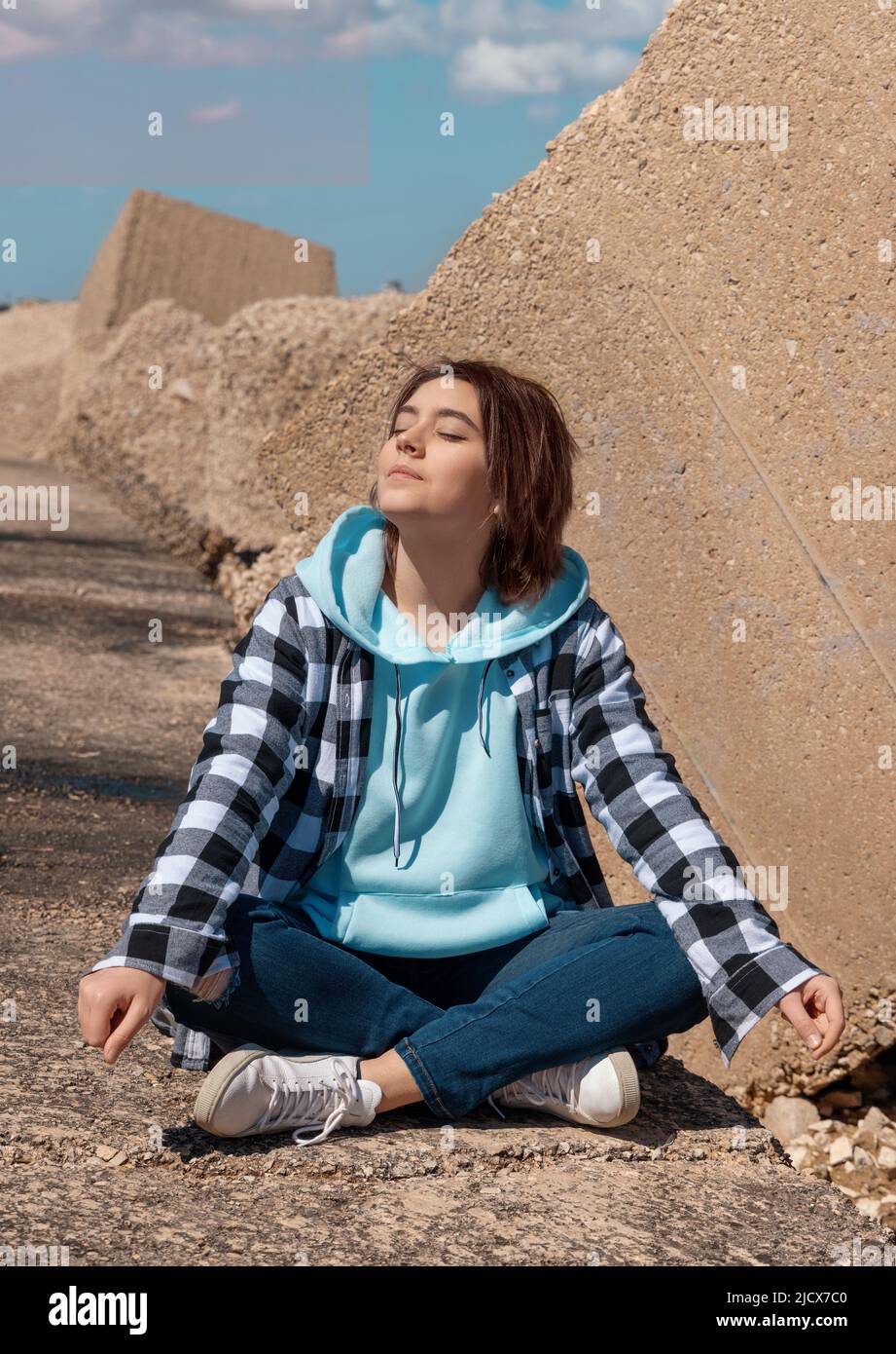 Happy Teenager girl in light blue hoodie and plaid shirt. Smiling Teen girl sitting outdoor cross-legged and enjoying the sunny day. Adolescence conce Stock Photo