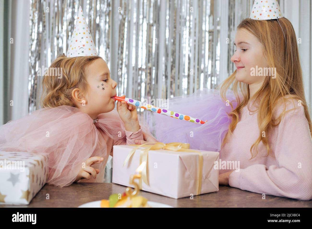 Two cute sisters celebrate birthday of youngest, who turned 5 years old. Girls having fun with festive horn, there are gift boxes and cake in front of Stock Photo