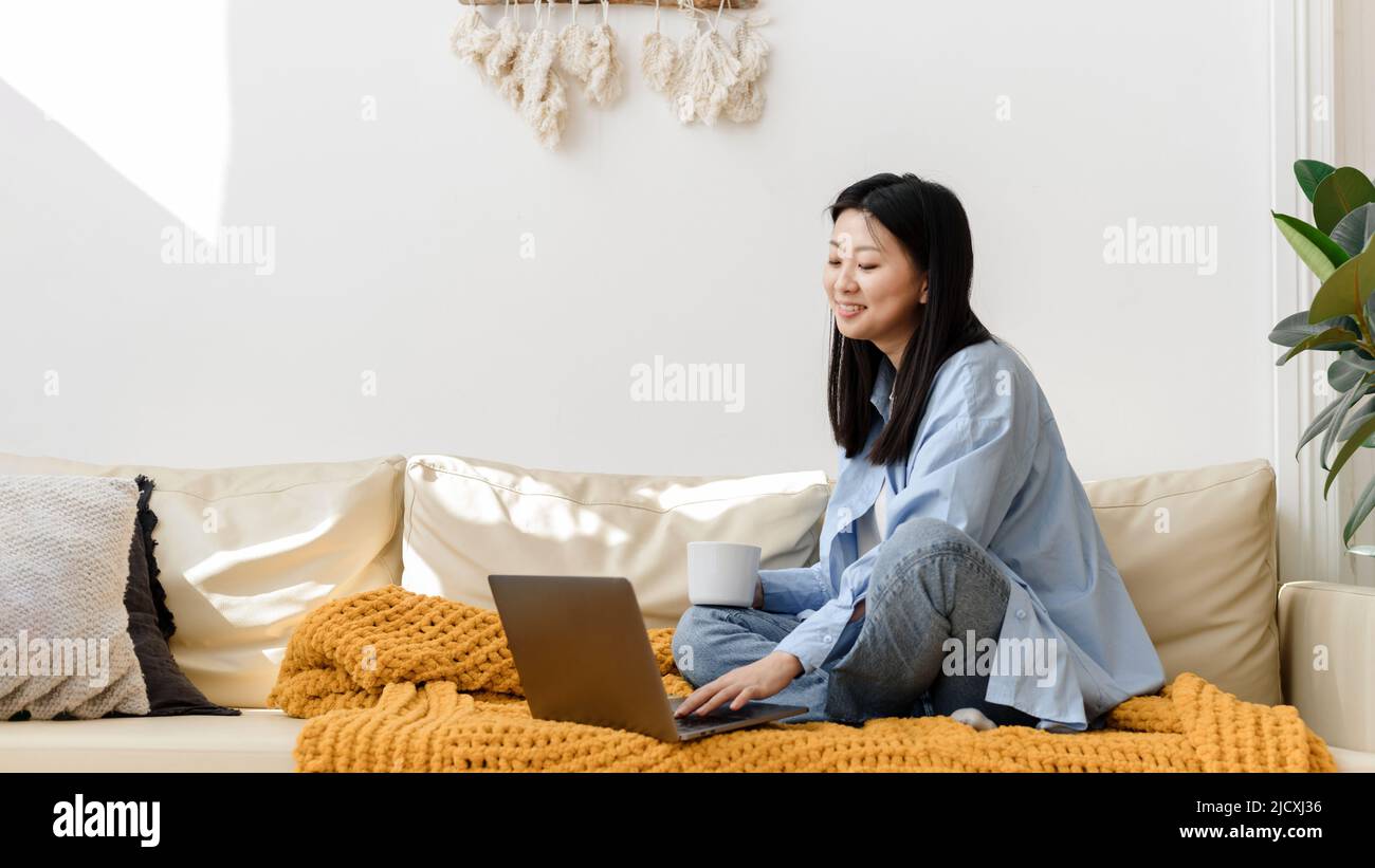 Portrait of smiling asian woman sitting on sofa using laptop in cozy ...