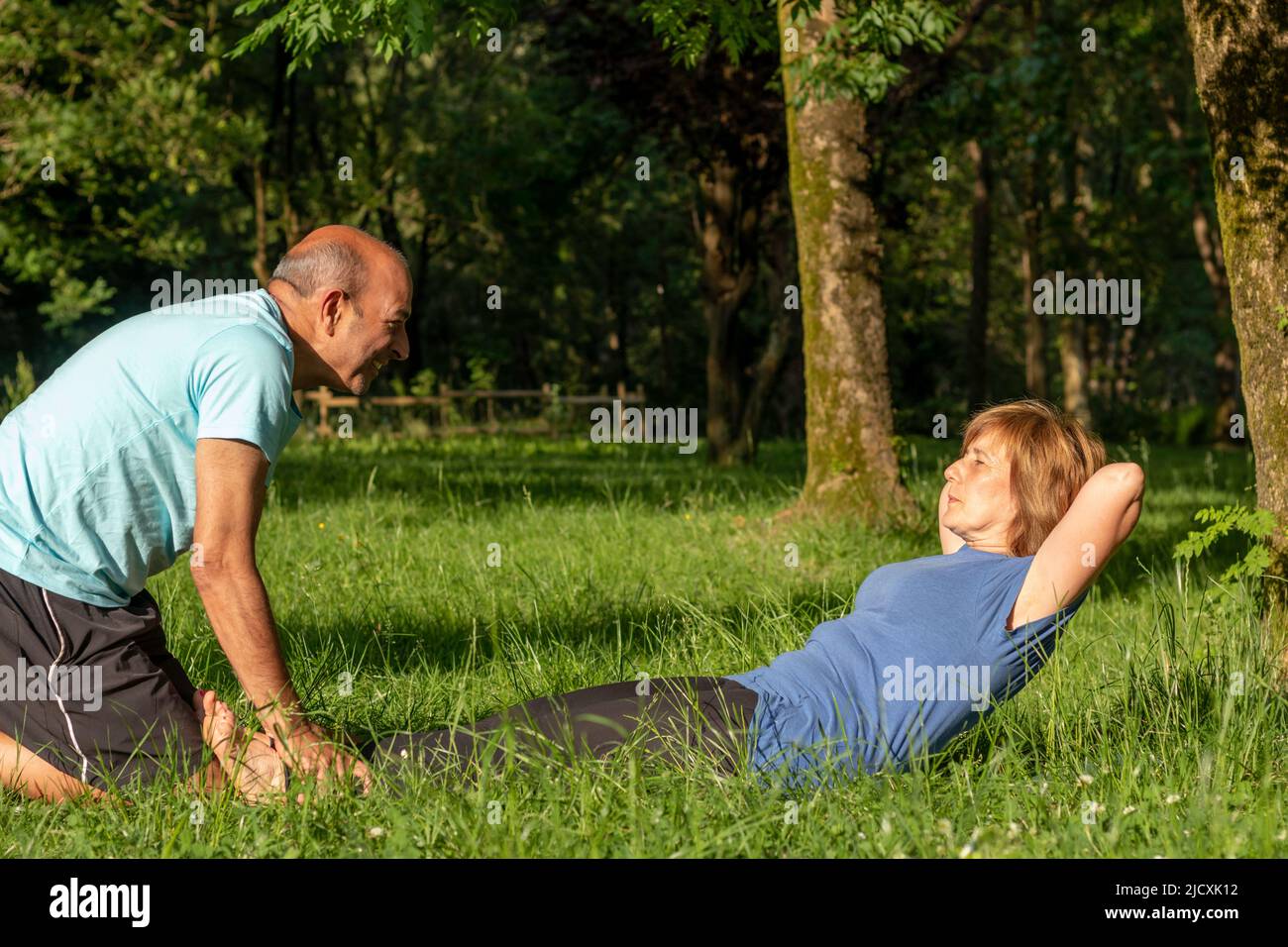 Woman trying to do some crunches in the park with her help of her husband in natural enviroment with trees behind Stock Photo