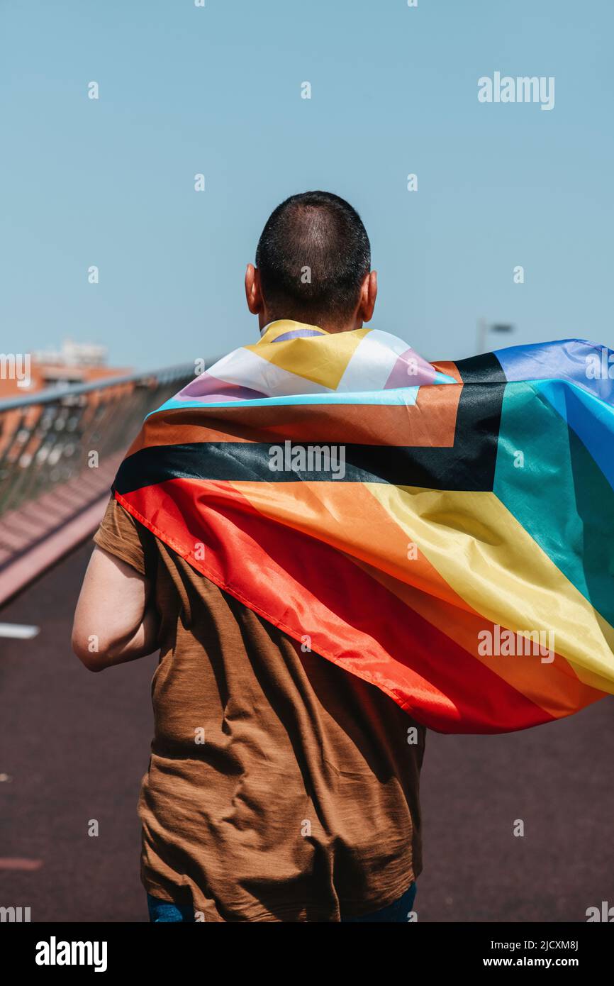 a man, seen from behind, wears an intersex-inclusive progress pride flag wrapped around his neck, walking by a bridge Stock Photo