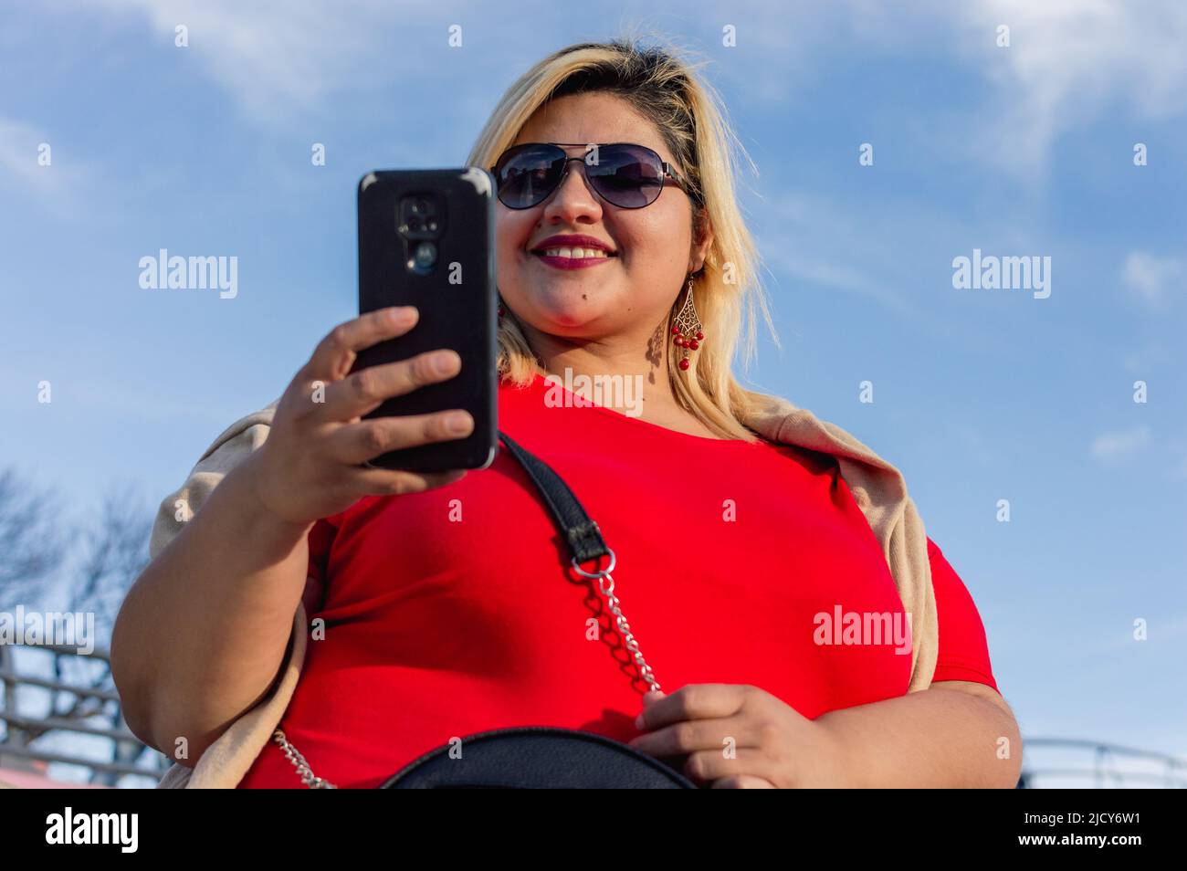 portrait cute young plus size caucasian latin woman dressed in red, smiling happy checking the phone in sending messages, standing outdoors with the s Stock Photo