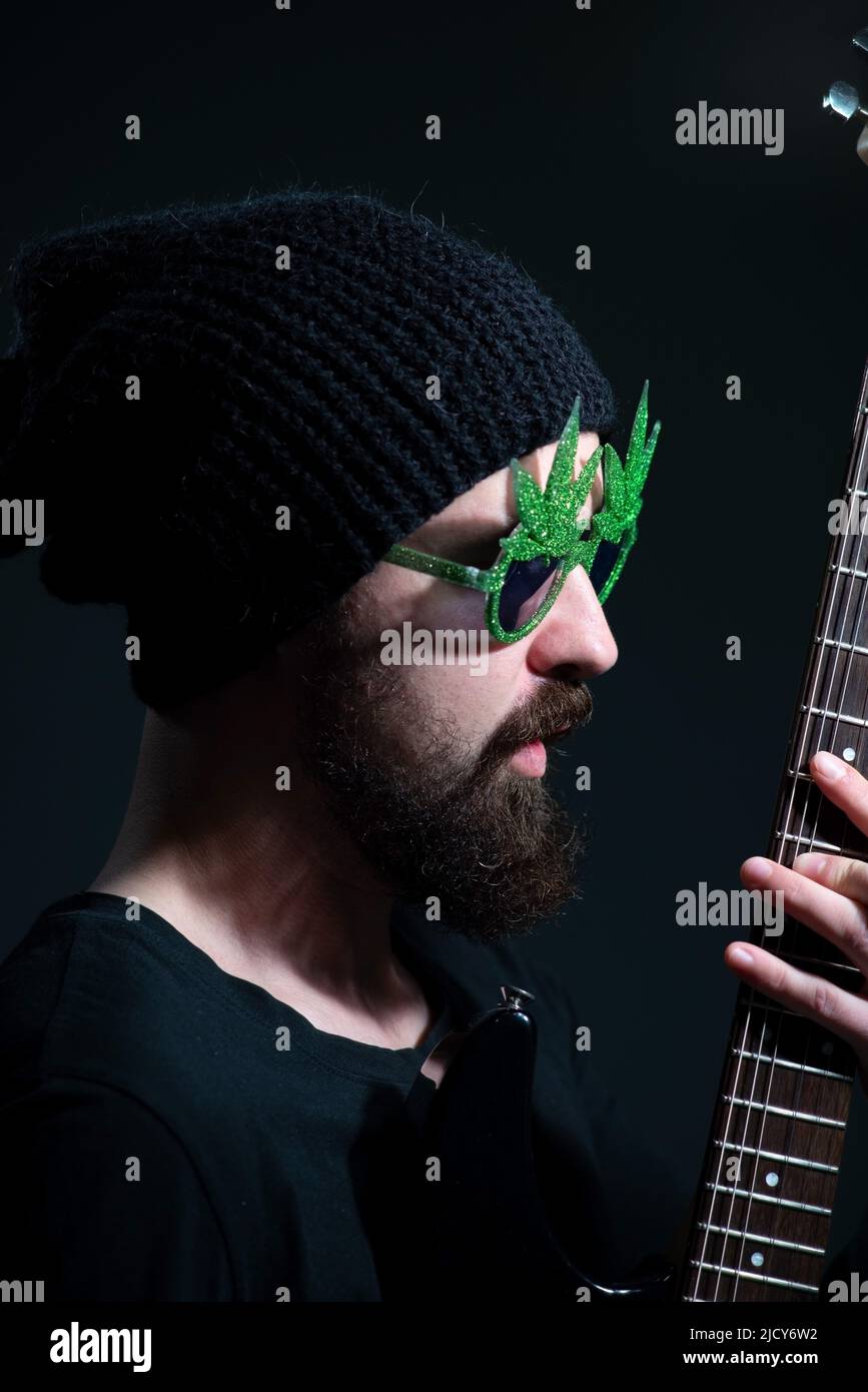 portrait of a bearded musician guy, wearing marijuana sunglasses, emotionally playing the electric guitar. on a dark background Stock Photo