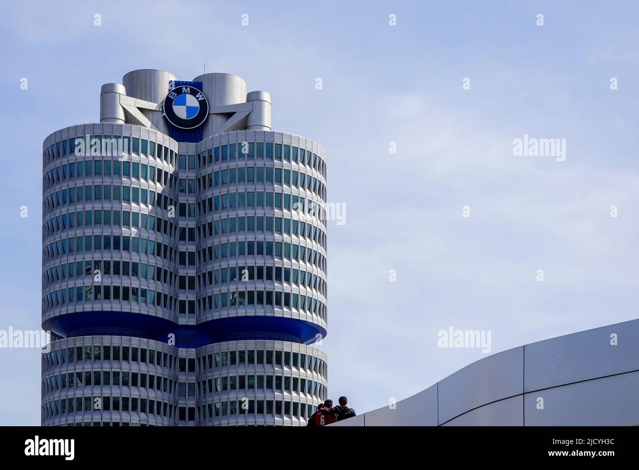 Three guys in front of the BMW Tower, the BMW four-cylinder is the administration building of the car manufacturer BMW in Munich, Germany, 12.4.22 Stock Photo