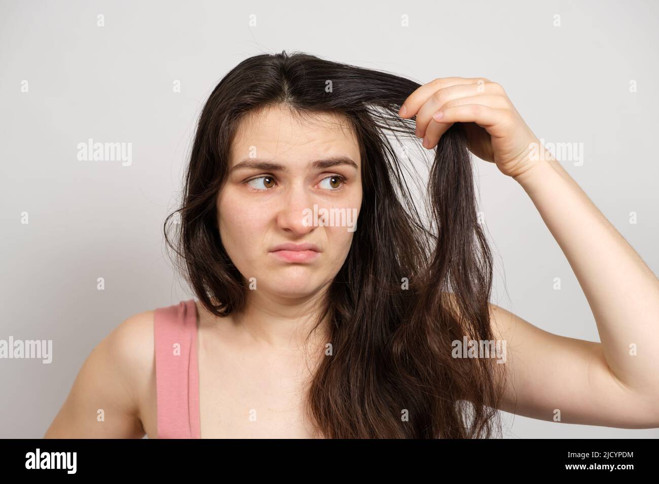 A beautiful brunette woman without makeup with dirty oily hair holds her hands on her head with a dissatisfied look Stock Photo