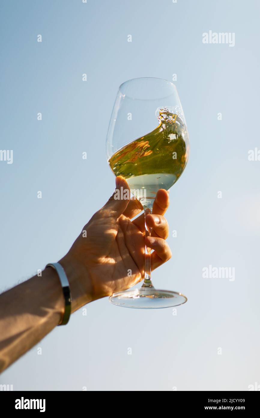Against the sky, a man holds a glass with wine, which is spinning in a glass Stock Photo