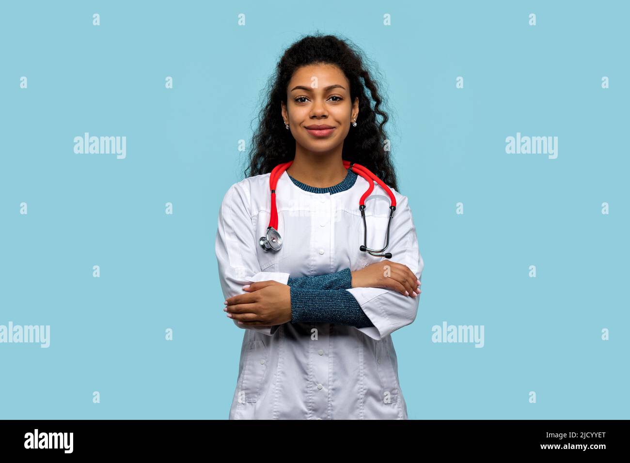 Portrait Positive Non-Caucasian Skinned Woman Doctor with crossed Arms Smiling Looking at Camera on Blue Studio Wall. African American Female Medical Stock Photo