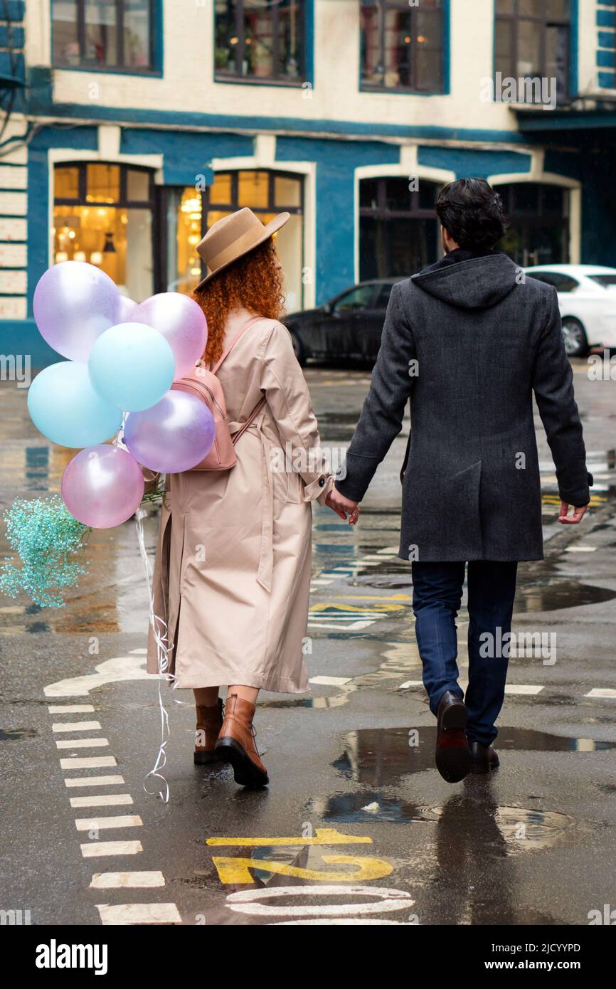 Heterosexual Couple on Romantic Date on Rainy Evening. Caucasian Lovers Walks City Streets with Bouquet Blue Flowers and Balloons. Girlfriend and boyf Stock Photo