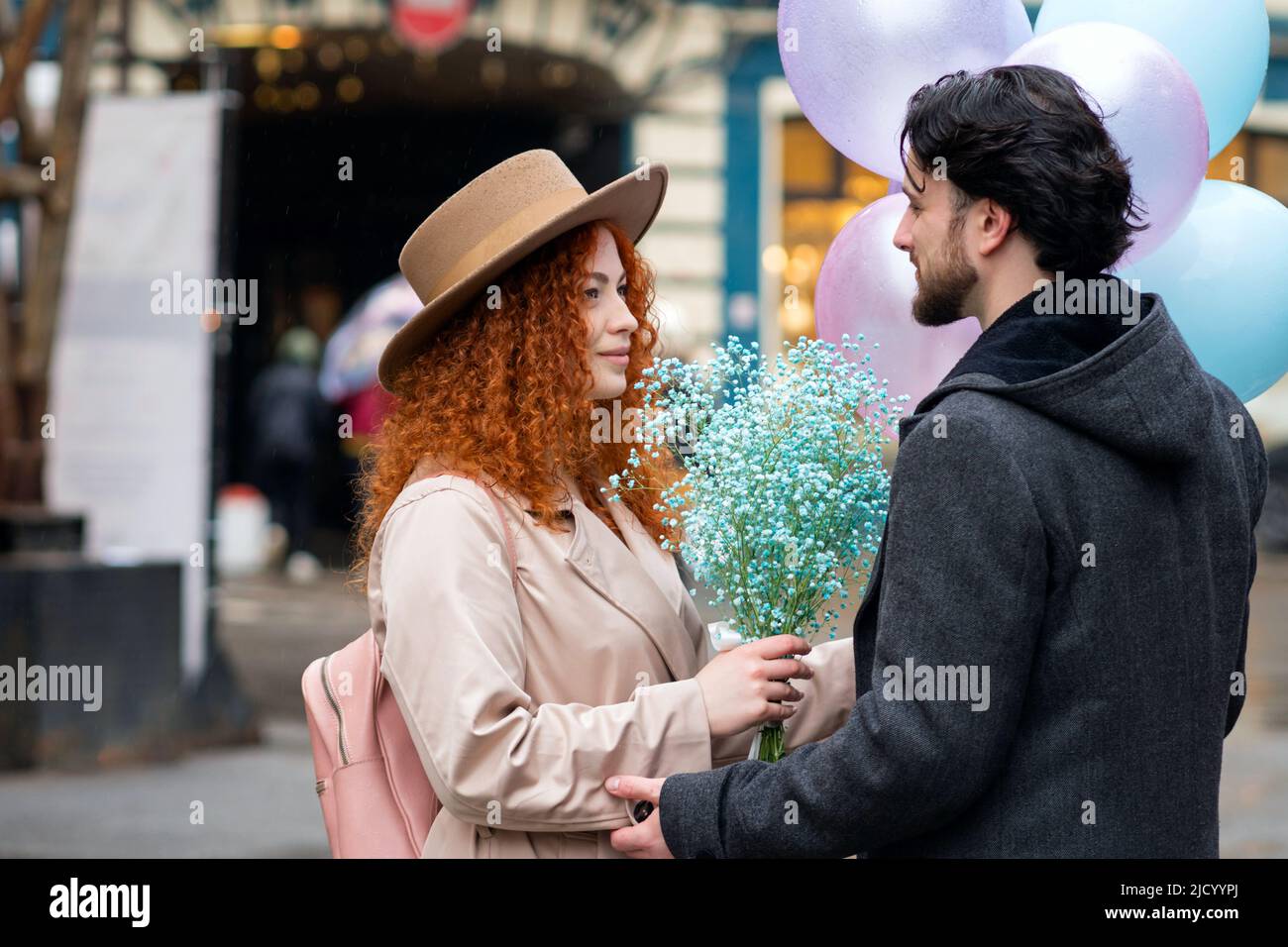 Heterosexual Couple in Love on Romantic Date on Rainy Evening. Caucasian Lovers Walks through City Streets with Bouquet Blue Flowers and Helium Balloo Stock Photo