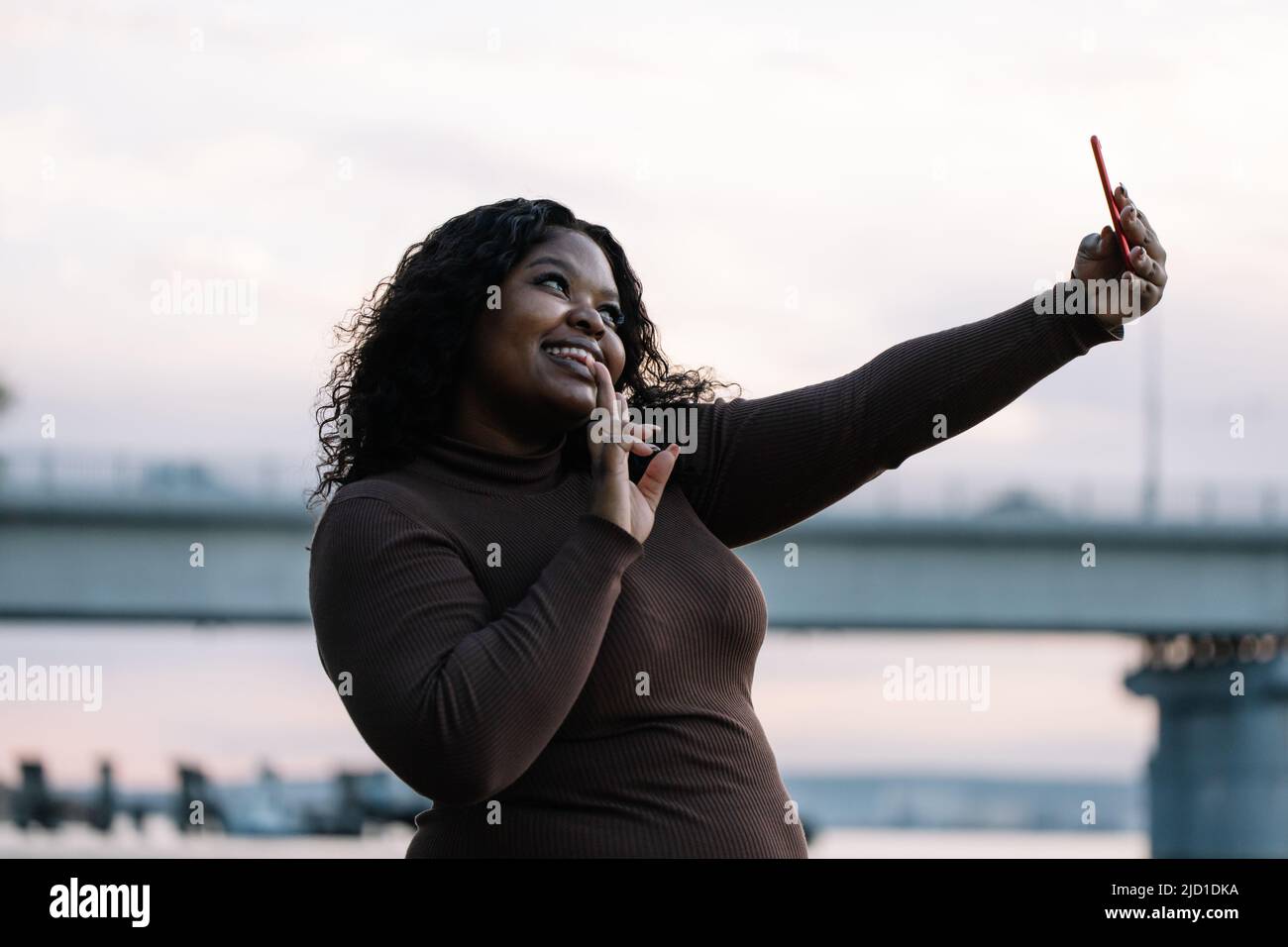 Curly african american plump woman in dress taking selfie, making duck face by smartphone, posing against sunset sky Stock Photo