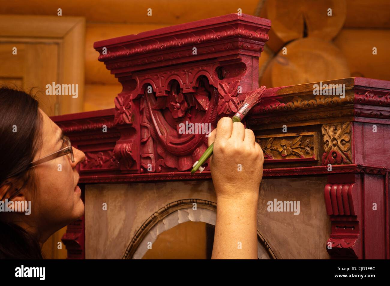 Attentive lady coloring massive wooden cupboard with astounding carved ornaments in red with brush with house walls in background. Reuse of old Stock Photo