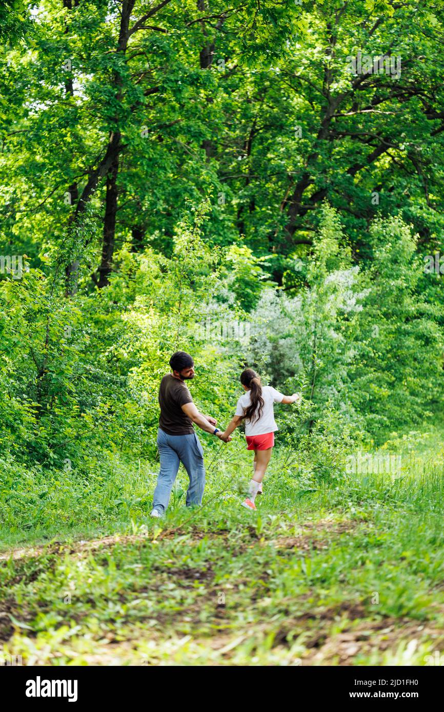 Back view of happy family walking in park forest around green trees, having fun. Little daughter holding hand of father. Stock Photo