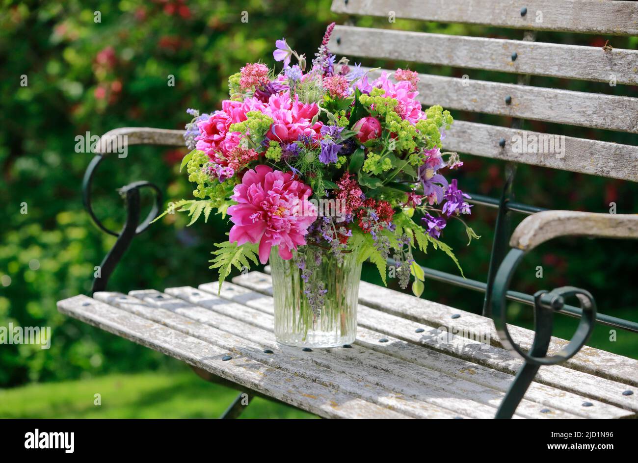 Colourful bouquet in red, pink and violet shades with peonies and columbines, stands in glass vase on decorative wooden garden bench Stock Photo