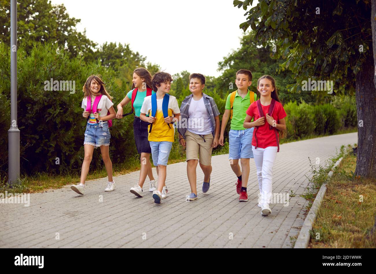 Funny schoolchildren group with backpacks have fun walking together on path in park. Stock Photo