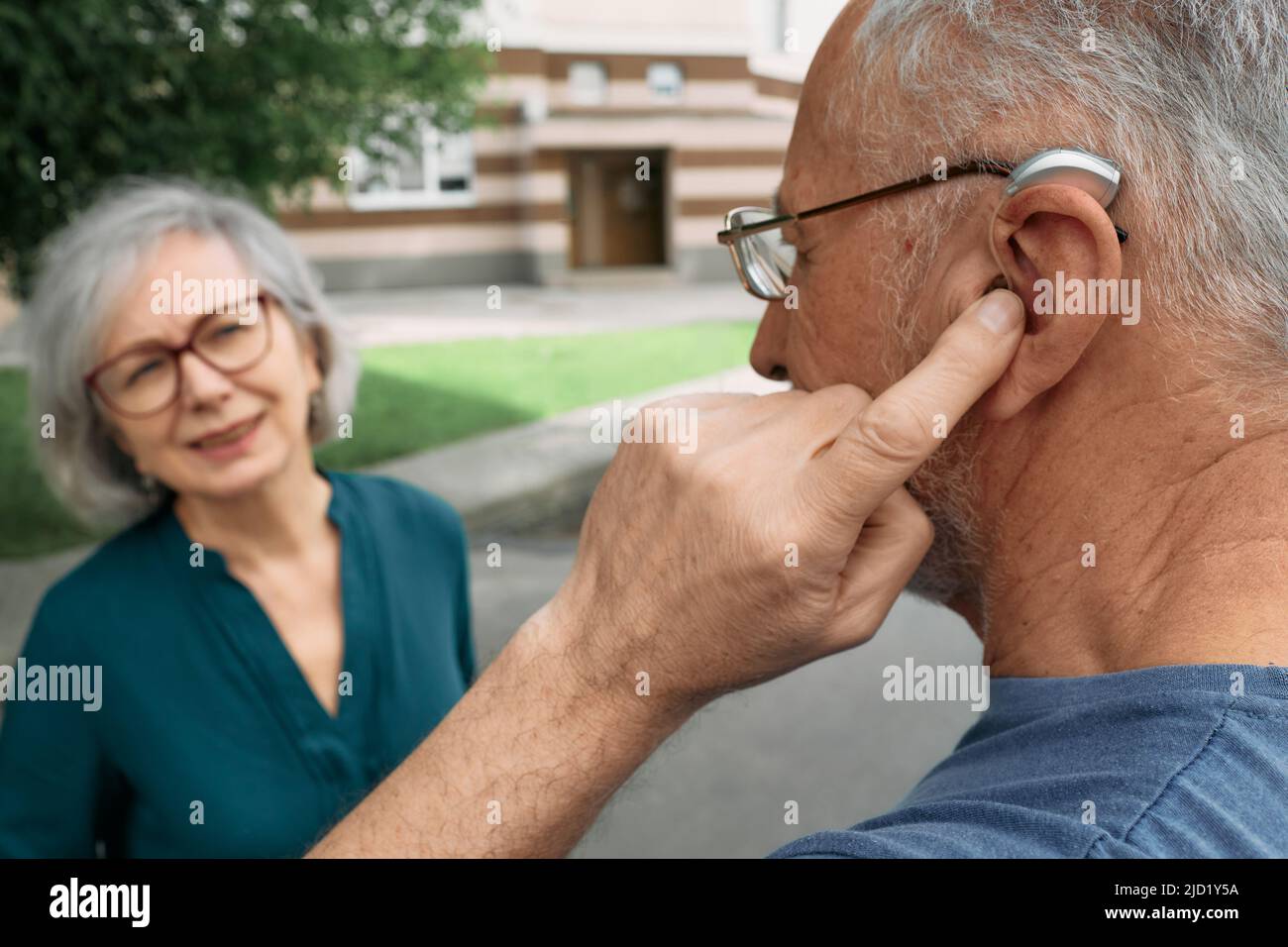 Mature man with a hearing impairment uses a hearing aid to communicate with his female senior friend outdoor. Hearing solutions Stock Photo