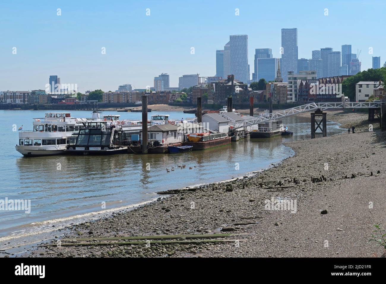 London, UK. River Thames sightseeing boats moored at a jetty at Bermondsey. Low tide reveals the foreshore. Towers of Canary Wharf in background. Stock Photo