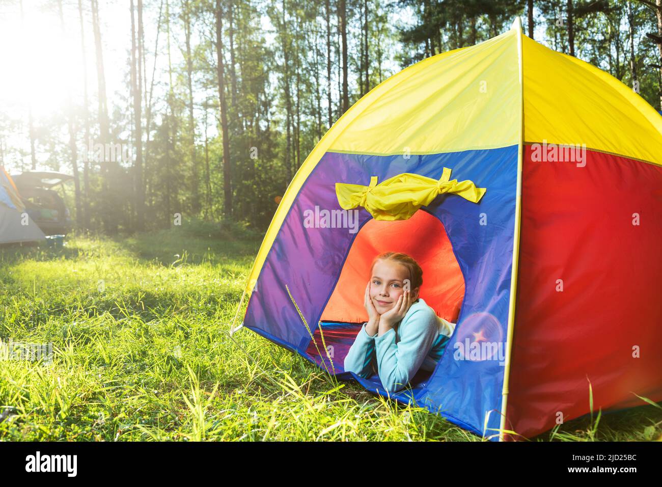 Little girl lying in a tent on a sunny forest meadow. The sun shines through the trees. The child looks at the camera, enjoying a sunny day. Family su Stock Photo