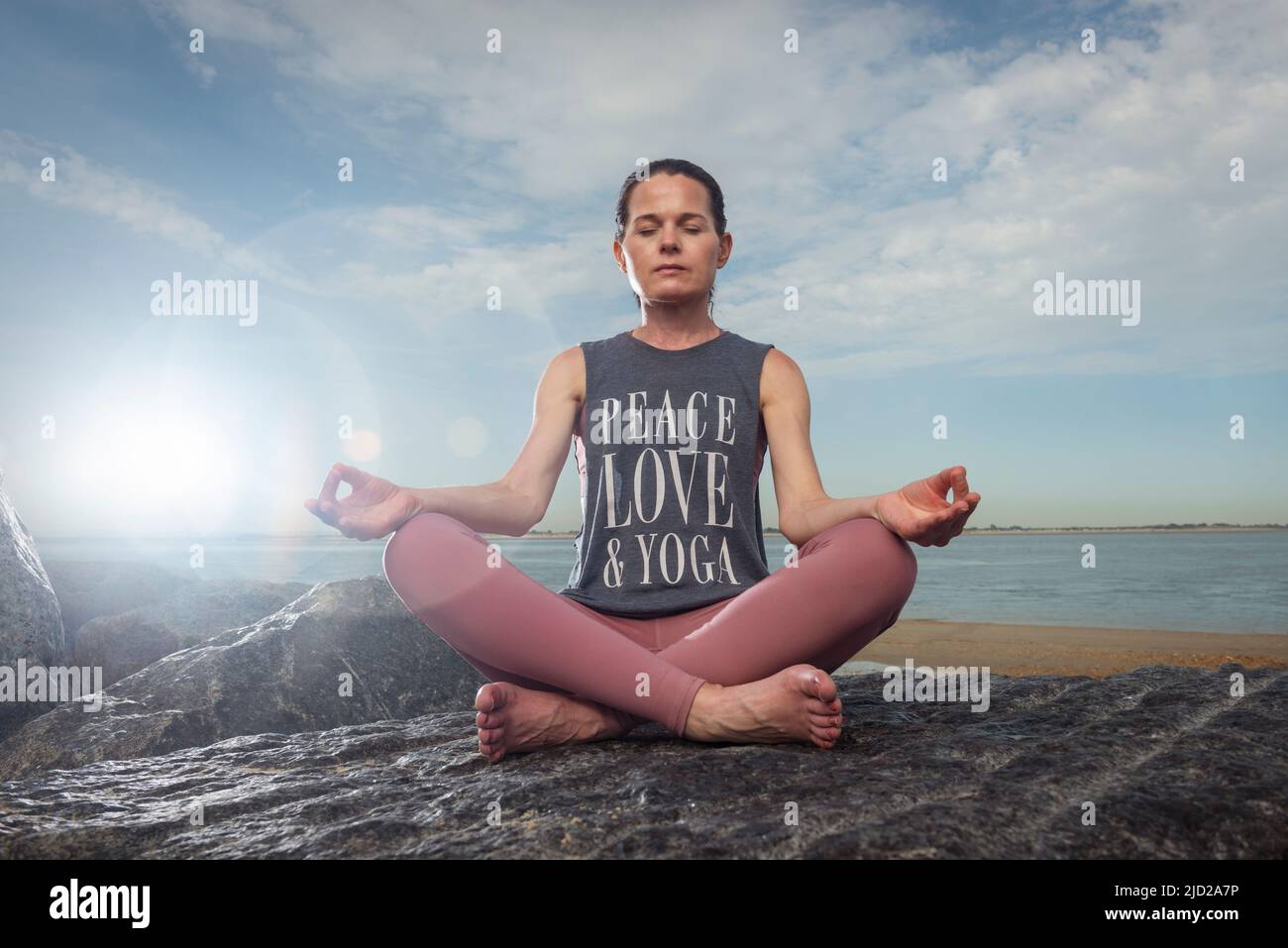 Woman sitting on rocks by the sea wearing a t shirt with 'peace love and yoga' slogan on it. Stock Photo