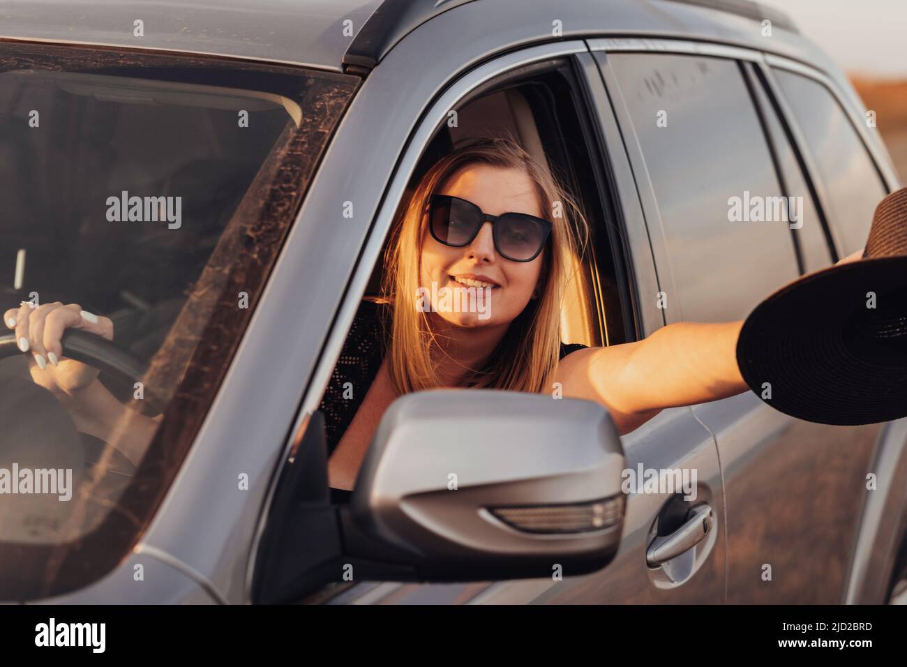 Cheerful Young Woman Driving Car and Showing the Hat in Hand Through Window at Sunset Stock Photo