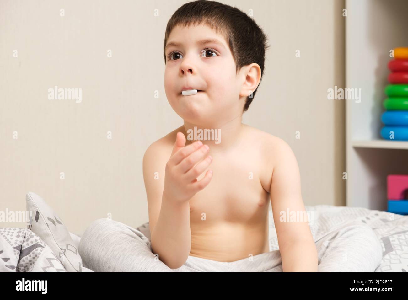 A 4yearold boy sits on a bed with a large white pill in his mouth