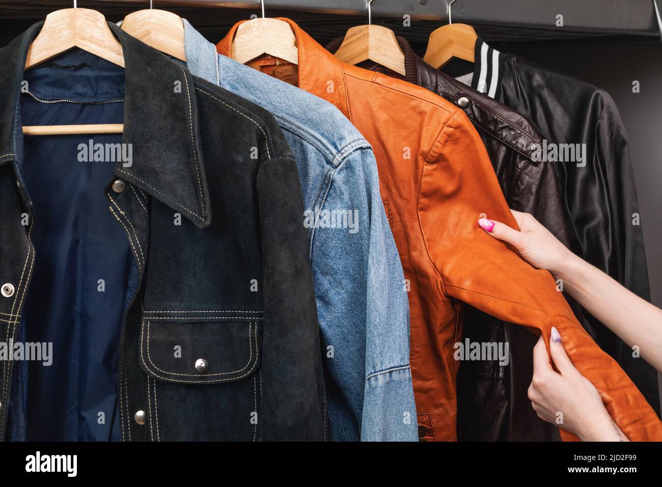 Woman choosing clothing in a second hand store. Various vintage suede leather and jeans jackets hang on clothing rack. Thrifting and sustainability in Stock Photo