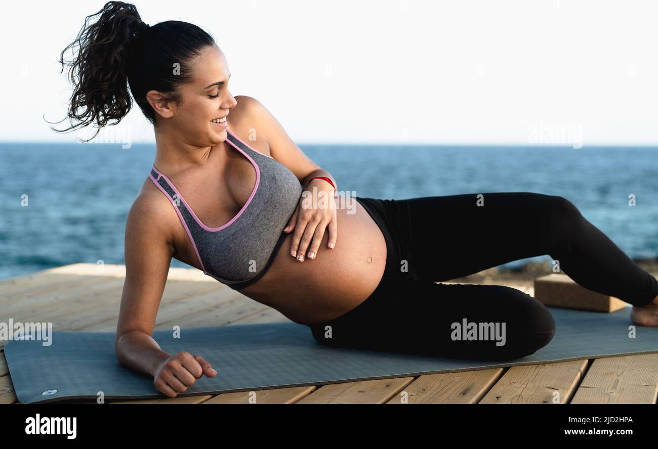Happy young woman caressing her pregnant belly while doing yoga next the ocean - Health lifestyle and maternity concept Stock Photo