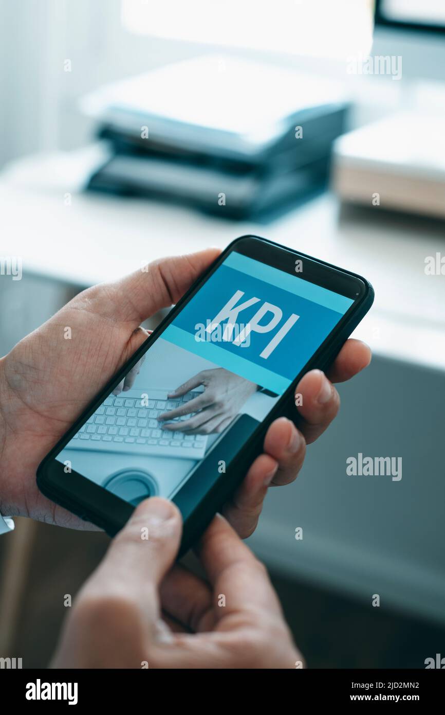a man, wearing a white shirt and a gray suit jacket, at the offic, holds his smartphone, that reads the text KPI, for key performance indicator, in it Stock Photo