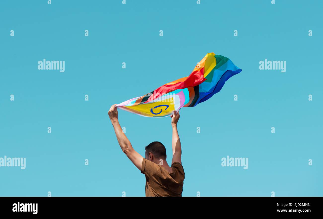 closeup of a man waving an intersex-inclusive progress pride flag above his head against the blue sky in a sunny day Stock Photo