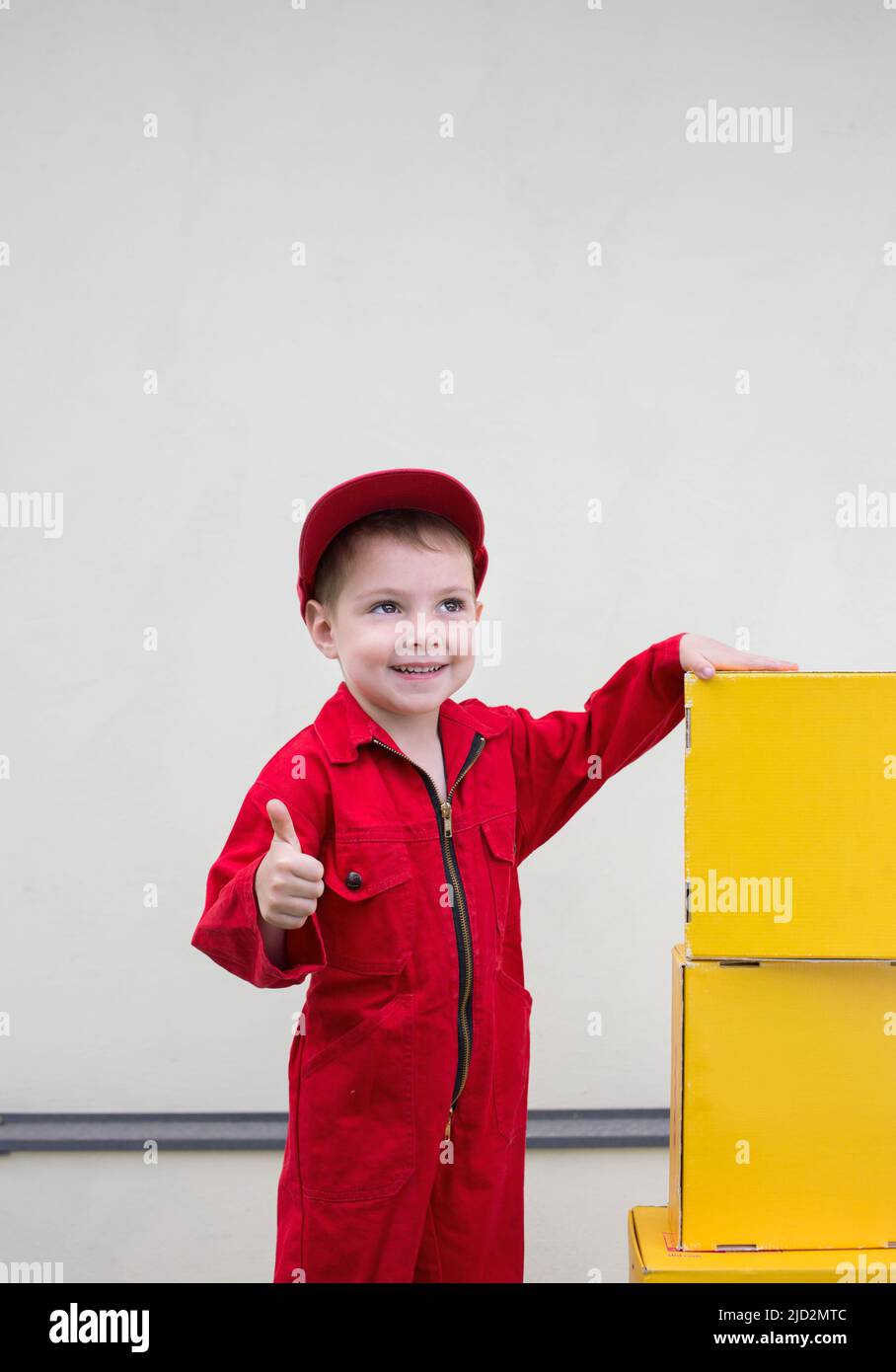 boy 3-4 years old in a red overalls, work uniform and a cap shows a like sign near yellow cardboard boxes. positive atmosphere. happiness delivery. Po Stock Photo