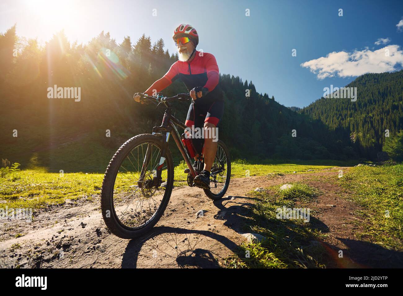 Old cyclist with white beard rides his mountain bike in the green spruce forest in red costume in Almaty, Kazakhstan. Extreme Sport and outdoor recrea Stock Photo
