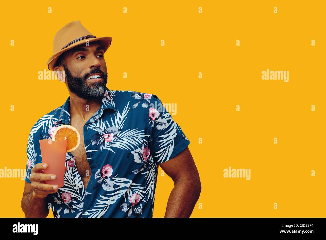 handsome bearded mid adult african american man wearing Hawaiian shirt and hat smiling with orange juice cocktail looking away studio Stock Photo