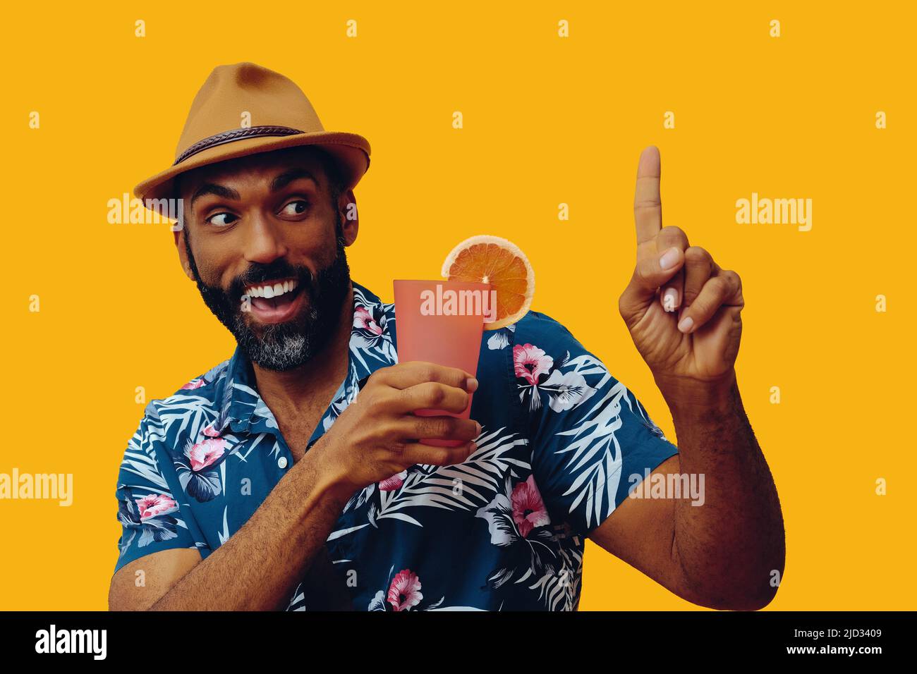 happy bearded mid adult african american man wearing Hawaiian shirt and hat smiling and orange juice cocktail pointing up studio Stock Photo