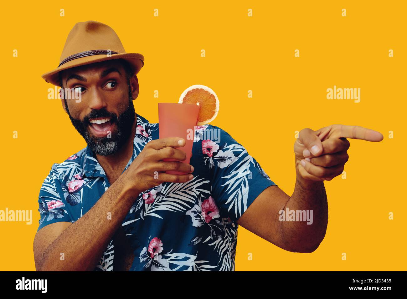 happy bearded mid adult african american man wearing Hawaiian shirt and hat smiling and orange juice cocktail pointing and looking away studio  Stock Photo