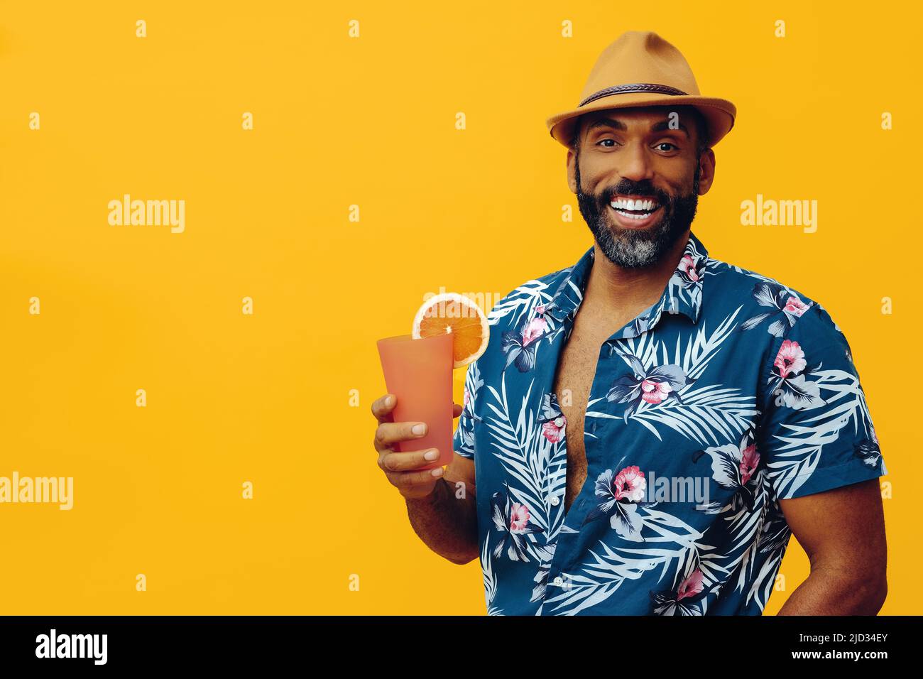 handsome bearded mid adult african american man wearing Hawaiian shirt and hat smiling with orange juice cocktail looking at camera studio Stock Photo