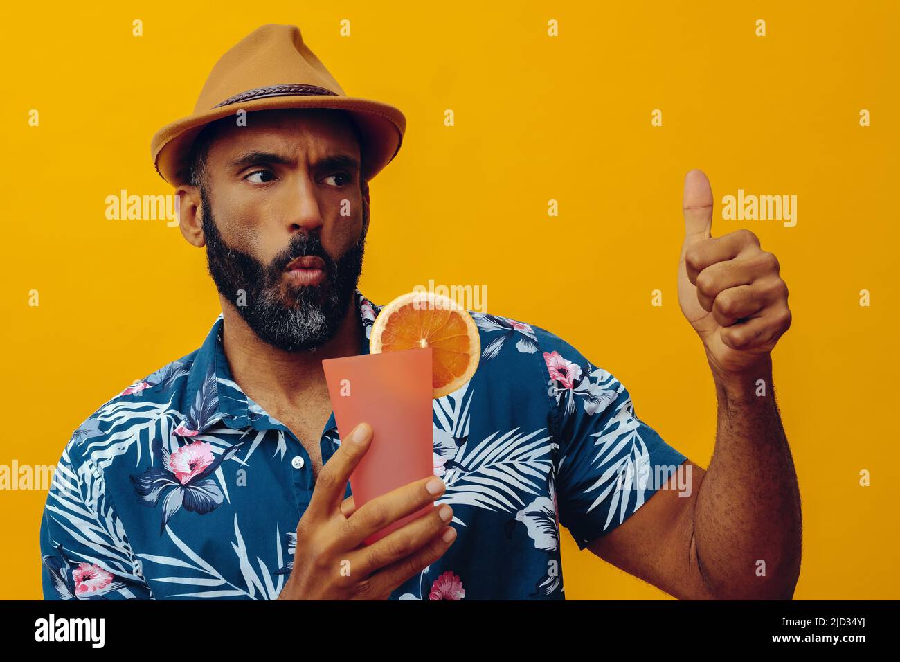 happy bearded mid adult african american man wearing Hawaiian shirt and hat with thumbs up and orange juice cocktail looking away studio Stock Photo